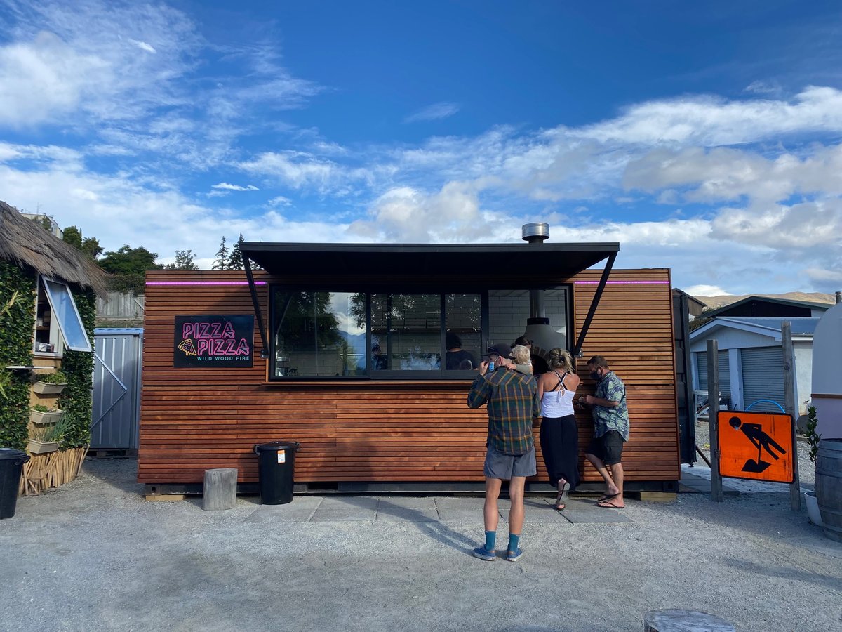 A wooden food truck with a serving window, customers lined up outside on a sunny day with blue sky