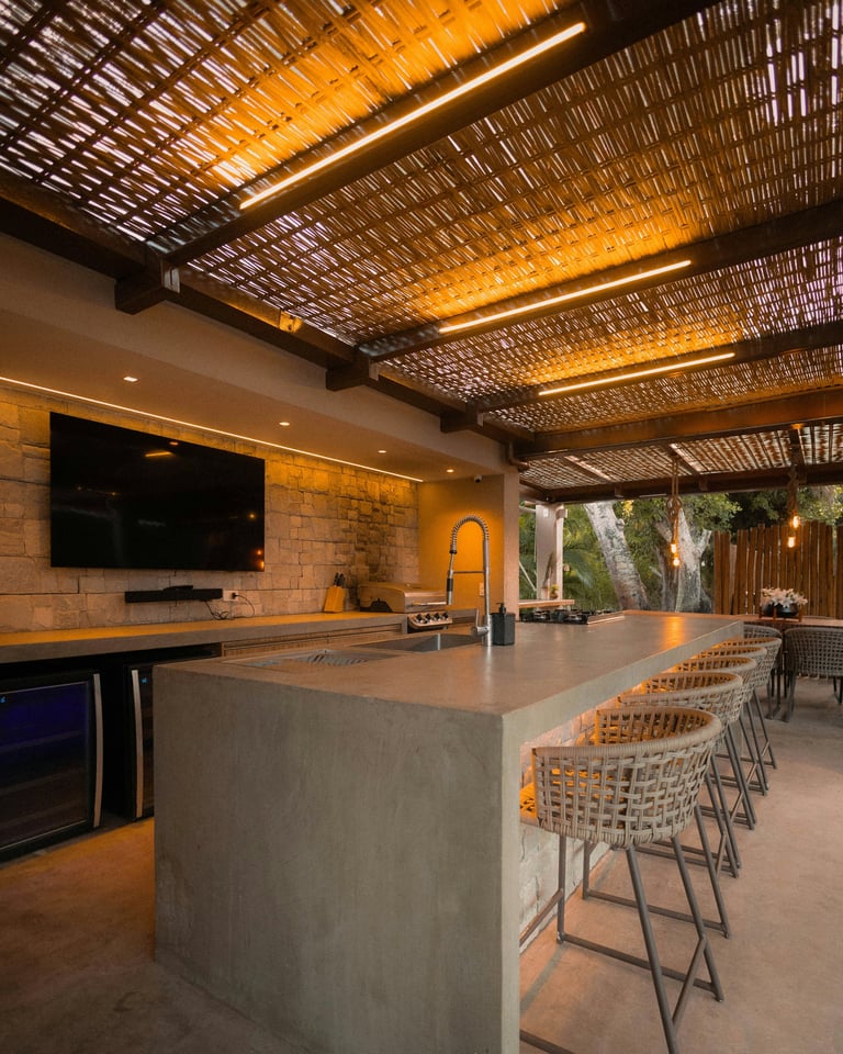 Modern outdoor kitchen with concrete island, rattan ceiling, warm lighting, and comfortable seating overlooking a backyard patio