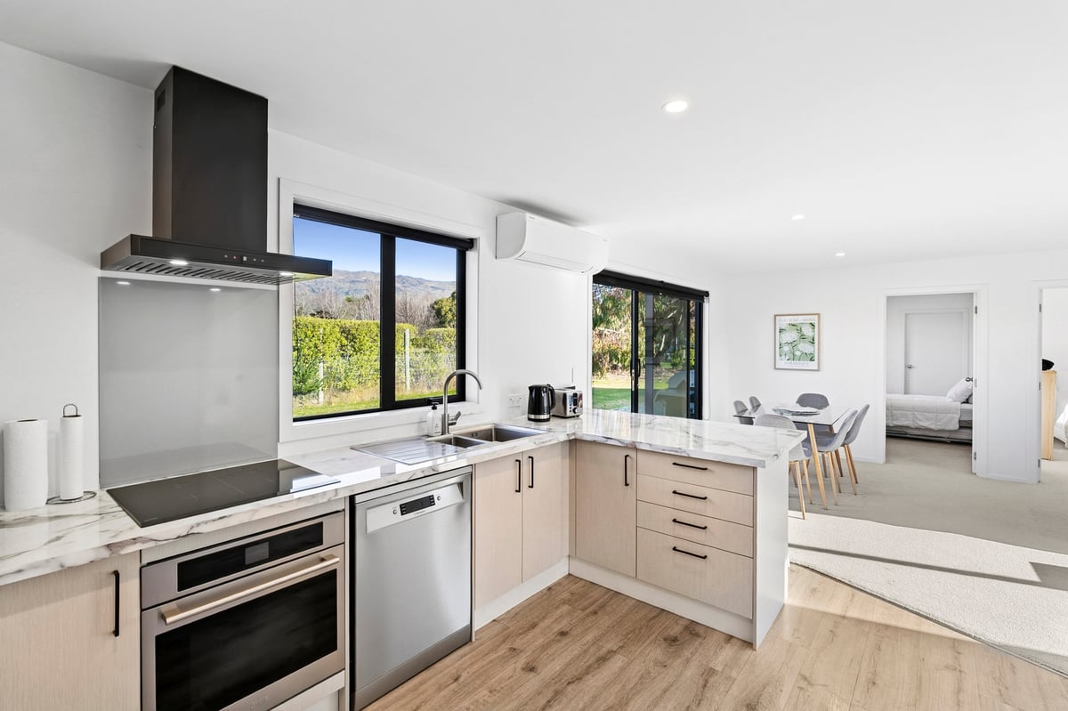 Modern kitchen with stainless steel appliances, light wood cabinetry, black range hood, and large windows overlooking greenery